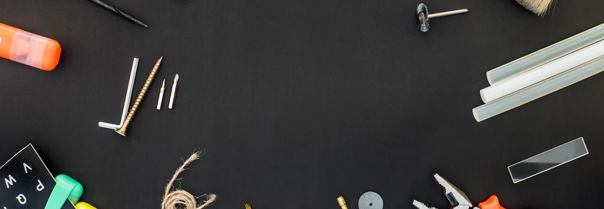 A variety of tools and items are arranged against a black background. These include a drill, a paintbrush, screws, drill bits, pliers, an eraser, some markers, a measuring tape, a spool of twine, and several other small hardware items.