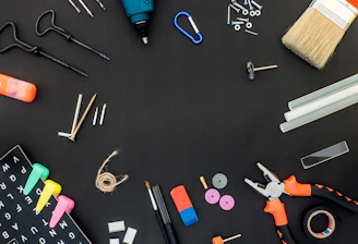 A variety of tools and items are arranged against a black background. These include a drill, a paintbrush, screws, drill bits, pliers, an eraser, some markers, a measuring tape, a spool of twine, and several other small hardware items.