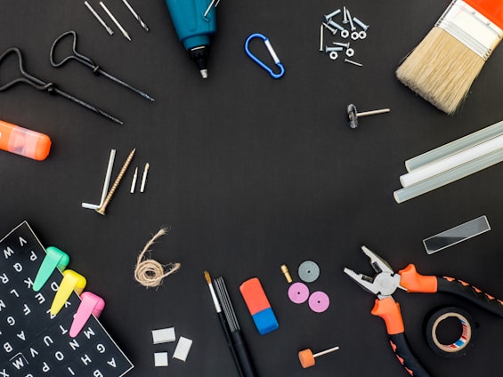 A variety of tools and items are arranged against a black background. These include a drill, a paintbrush, screws, drill bits, pliers, an eraser, some markers, a measuring tape, a spool of twine, and several other small hardware items.