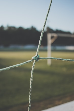 A close-up of a knot in a green soccer goal net, with a blurry background featuring a grassy field and goalposts.