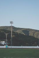 A sports field with green grass is bordered by a running track and enclosed by a white wall. Beyond the field, there are two tall light towers and several small buildings. The background features lush green hills under a clear blue sky.