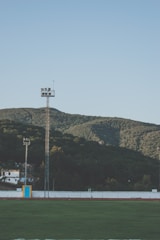 A sports field with green grass is bordered by a running track and enclosed by a white wall. Beyond the field, there are two tall light towers and several small buildings. The background features lush green hills under a clear blue sky.