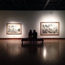 A dimly lit art gallery features two large framed paintings on the wall, showcasing landscape scenes with mountains and mist. In the foreground, two silhouetted figures sit closely on a bench, facing the art pieces.