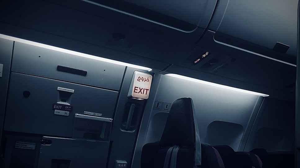An airplane interior showing the exit area, with overhead compartments and signage in both English and another language. The seating area is visible with dim ambient lighting, giving a calm atmosphere.