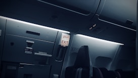 An airplane interior showing the exit area, with overhead compartments and signage in both English and another language. The seating area is visible with dim ambient lighting, giving a calm atmosphere.