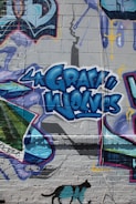 Colorful graffiti artwork on a brick wall, featuring the words 'Gray Wolves' in bold blue letters. The mural is filled with vibrant shapes and patterns, with a shadow stencil of a spray can above. A silhouette of a black cat with teal patches is shown at the bottom.
