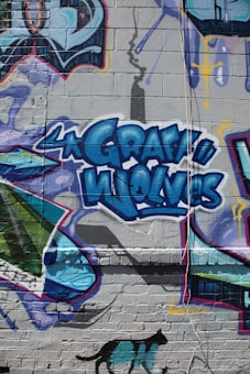 Colorful graffiti artwork on a brick wall, featuring the words 'Gray Wolves' in bold blue letters. The mural is filled with vibrant shapes and patterns, with a shadow stencil of a spray can above. A silhouette of a black cat with teal patches is shown at the bottom.