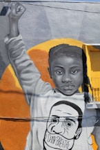 A mural depicts a young African American girl raising her fist in a gesture of power and solidarity. Her facial expression is serious and resolute. She has long braids and is wearing a sweatshirt that bears the face of a man with the words 'I CAN'T BREATHE' written across his mask. The background is an orange circle that contrasts with the monochrome tones of the mural.