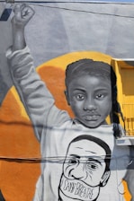A mural depicts a young African American girl raising her fist in a gesture of power and solidarity. Her facial expression is serious and resolute. She has long braids and is wearing a sweatshirt that bears the face of a man with the words 'I CAN'T BREATHE' written across his mask. The background is an orange circle that contrasts with the monochrome tones of the mural.