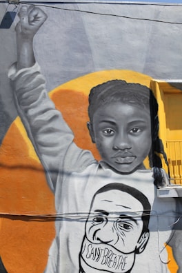 A mural depicts a young African American girl raising her fist in a gesture of power and solidarity. Her facial expression is serious and resolute. She has long braids and is wearing a sweatshirt that bears the face of a man with the words 'I CAN'T BREATHE' written across his mask. The background is an orange circle that contrasts with the monochrome tones of the mural.
