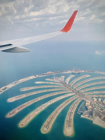 An aerial view of a man-made island shaped like a palm tree, surrounded by clear blue water. The image captures the intricate design of the island's fronds lined with buildings and roads. A part of an airplane wing is visible in the top left corner, suggesting the photo was taken from a plane. The sky is partly cloudy, contributing to a serene and expansive feel.
