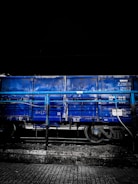 A blue freight train car is stationed on railway tracks. The car is partially obscured by a blue railing, and the image has a dark, moody background, suggesting it may be nighttime. The ground consists of a cobblestone or tiled surface.