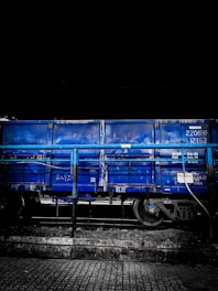 A blue freight train car is stationed on railway tracks. The car is partially obscured by a blue railing, and the image has a dark, moody background, suggesting it may be nighttime. The ground consists of a cobblestone or tiled surface.