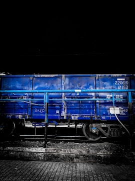 A blue freight train car is stationed on railway tracks. The car is partially obscured by a blue railing, and the image has a dark, moody background, suggesting it may be nighttime. The ground consists of a cobblestone or tiled surface.