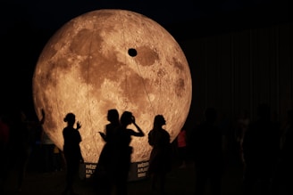 Silhouetted figures stand in front of a large, illuminated model of the moon, featuring detailed lunar craters and surface textures. The scene is set in a dimly lit environment, highlighting the contrast between the bright model and the dark surroundings.