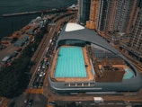 An aerial view captures an urban swimming pool complex surrounded by high-rise buildings. The pool is rectangular and features multiple swimming lanes, with a few umbrellas providing shade near the edges. Adjacent roads are lined with vehicles, suggesting a bustling city environment. The complex is located near a waterfront with various cargo containers and cranes visible in the distance.