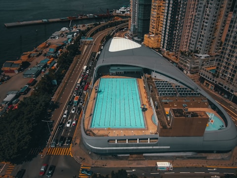 An aerial view captures an urban swimming pool complex surrounded by high-rise buildings. The pool is rectangular and features multiple swimming lanes, with a few umbrellas providing shade near the edges. Adjacent roads are lined with vehicles, suggesting a bustling city environment. The complex is located near a waterfront with various cargo containers and cranes visible in the distance.