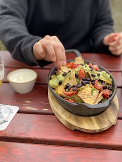 A person wearing a dark sweater is reaching for a serving of nachos topped with guacamole, tomatoes, and black beans, served in a cast iron skillet placed on a wooden board. The table is made of red wood, and a small bowl of sauce along with a beer glass is visible nearby.