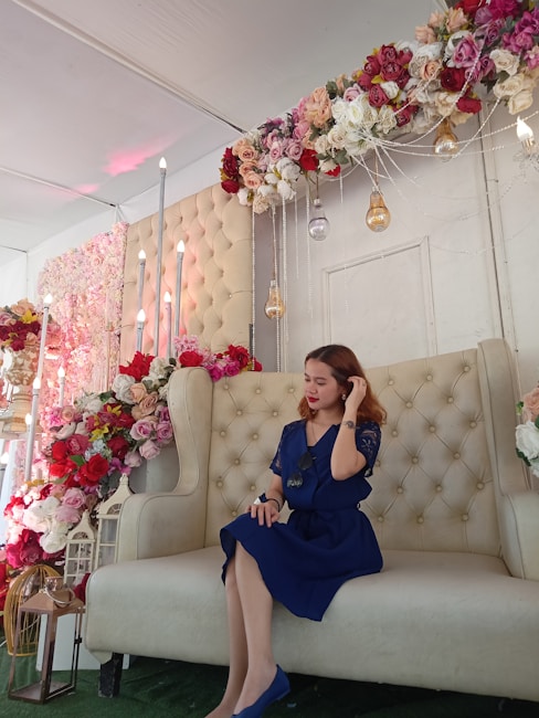 A woman in a blue dress sits on an elegant, cream-colored tufted sofa. The setting is adorned with abundant floral decorations, featuring red, pink, and white roses overhead and on the side. Tall candles and hanging decorative light bulbs enhance the ornate backdrop.