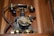 An ornate, vintage rotary dial telephone with a detailed metal base featuring engraved patterns. It has a metal rotary dial with numbered finger holes, a black handset resting on a cradle, and a curled cord connecting the handset to the base. The device is set against a wooden backdrop.