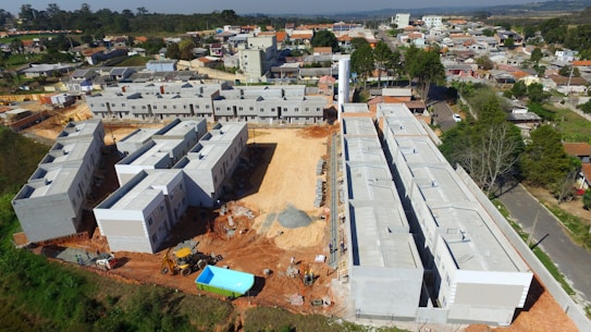 A construction site with several large buildings in various stages of completion, surrounded by dirt and construction equipment. The area is near a residential neighborhood with houses and trees visible in the background.