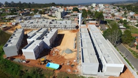 A construction site with several large buildings in various stages of completion, surrounded by dirt and construction equipment. The area is near a residential neighborhood with houses and trees visible in the background.