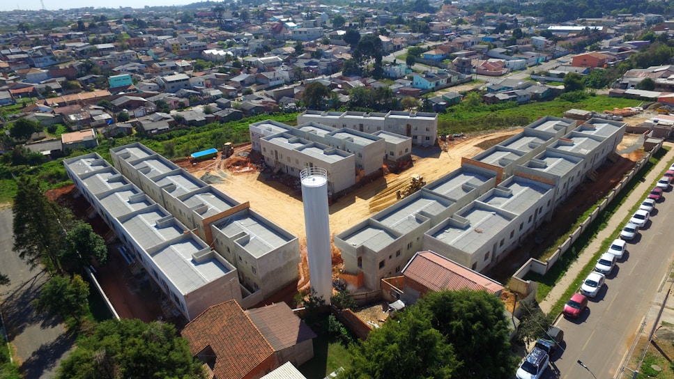 Aerial view of a residential construction site consisting of multiple gray buildings arranged in a structured manner. Surrounding the construction are numerous houses that form a densely packed urban neighborhood. There is a central cylindrical structure within the construction site, possibly a water tower. Various vehicles are parked along a road adjacent to the site, and greenery can be seen in the nearby areas.