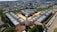 Aerial view of a residential construction site consisting of multiple gray buildings arranged in a structured manner. Surrounding the construction are numerous houses that form a densely packed urban neighborhood. There is a central cylindrical structure within the construction site, possibly a water tower. Various vehicles are parked along a road adjacent to the site, and greenery can be seen in the nearby areas.