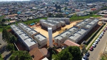 Aerial view of a residential construction site consisting of multiple gray buildings arranged in a structured manner. Surrounding the construction are numerous houses that form a densely packed urban neighborhood. There is a central cylindrical structure within the construction site, possibly a water tower. Various vehicles are parked along a road adjacent to the site, and greenery can be seen in the nearby areas.
