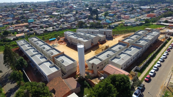 Aerial view of a residential construction site consisting of multiple gray buildings arranged in a structured manner. Surrounding the construction are numerous houses that form a densely packed urban neighborhood. There is a central cylindrical structure within the construction site, possibly a water tower. Various vehicles are parked along a road adjacent to the site, and greenery can be seen in the nearby areas.