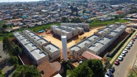 Aerial view of a residential construction site consisting of multiple gray buildings arranged in a structured manner. Surrounding the construction are numerous houses that form a densely packed urban neighborhood. There is a central cylindrical structure within the construction site, possibly a water tower. Various vehicles are parked along a road adjacent to the site, and greenery can be seen in the nearby areas.