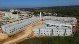 A construction site features multiple rows of newly built, unpainted apartment buildings made of concrete. Heavy machinery, including excavators, is visible around the site. The backdrop includes a suburban area with residential homes and a forested area extending into the distance.