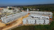 A construction site features multiple rows of newly built, unpainted apartment buildings made of concrete. Heavy machinery, including excavators, is visible around the site. The backdrop includes a suburban area with residential homes and a forested area extending into the distance.