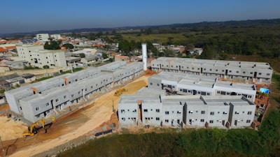 A construction site features multiple rows of newly built, unpainted apartment buildings made of concrete. Heavy machinery, including excavators, is visible around the site. The backdrop includes a suburban area with residential homes and a forested area extending into the distance.
