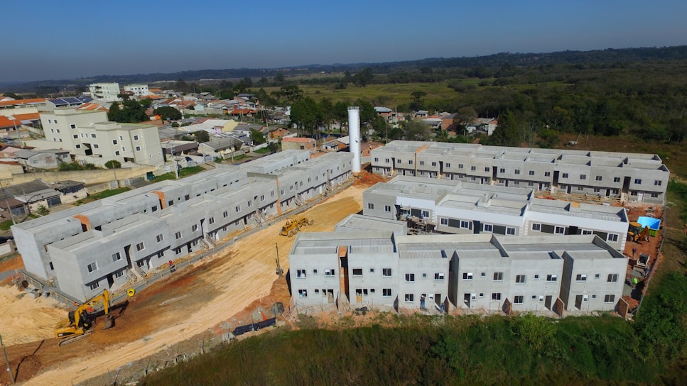 A construction site features multiple rows of newly built, unpainted apartment buildings made of concrete. Heavy machinery, including excavators, is visible around the site. The backdrop includes a suburban area with residential homes and a forested area extending into the distance.