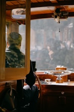 A person is seated at the helm of a wooden boat, surrounded by nautical equipment. The area features rich wood tones and a large steering wheel. The window reveals a hazy, indistinct view outside.