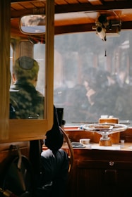 A person is seated at the helm of a wooden boat, surrounded by nautical equipment. The area features rich wood tones and a large steering wheel. The window reveals a hazy, indistinct view outside.