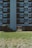 A several-story residential building with a predominantly brick facade featuring a central concrete section. The windows are arranged symmetrically, with some open and others closed, and balconies can be seen with various items. The bottom part of the building has an open passageway leading to another area. A grassy field is in the foreground, providing a green contrast to the building's darker tones.