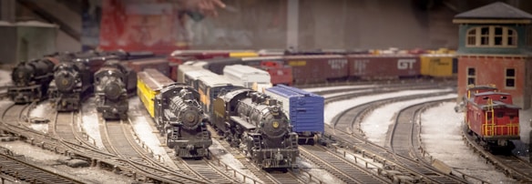 A detailed model train setup featuring multiple model trains on intersecting tracks with various types of cars, including steam locomotives and cargo cars. A small red building is situated to one side, and an assortment of freight cars is visible in the background.