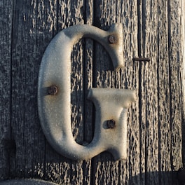 A large, metallic letter 'G' is mounted on a weathered wooden surface, displaying noticeable signs of rust around the attachment points.