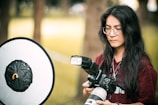 A person with long dark hair and glasses is holding a professional camera with a large lens, standing in an outdoor setting. A photography reflector is positioned nearby, indicating a photoshoot. The background shows blurred trees and greenery.