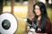 A person with long dark hair and glasses is holding a professional camera with a large lens, standing in an outdoor setting. A photography reflector is positioned nearby, indicating a photoshoot. The background shows blurred trees and greenery.