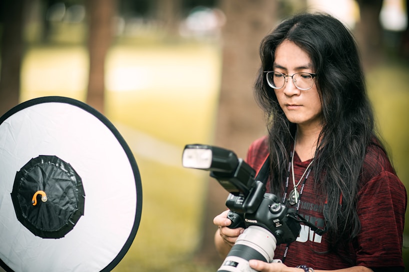 A person with long dark hair and glasses is holding a professional camera with a large lens, standing in an outdoor setting. A photography reflector is positioned nearby, indicating a photoshoot. The background shows blurred trees and greenery.