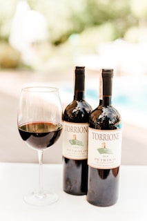 A close-up view of two bottles of wine labeled 'Torrione Petrolo 2015' and a glass filled with red wine. The background appears to be an outdoor setting with blurred greenery and a swimming pool.