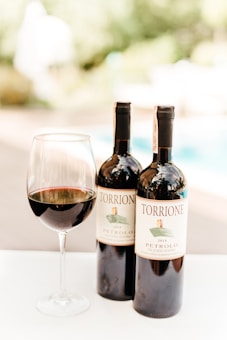 A close-up view of two bottles of wine labeled 'Torrione Petrolo 2015' and a glass filled with red wine. The background appears to be an outdoor setting with blurred greenery and a swimming pool.