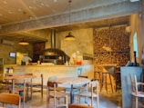 A warmly lit pizzeria with a rustic, industrial design featuring a large wood-fired oven. Stacks of firewood are stored on shelves along the walls. Round wooden tables and chairs are positioned throughout the room. Two chefs are working behind the counter, and there are a few condiments and utensils on top. The concrete floor and ceiling along with pendant lighting add to the cozy atmosphere.