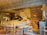 A warmly lit pizzeria with a rustic, industrial design featuring a large wood-fired oven. Stacks of firewood are stored on shelves along the walls. Round wooden tables and chairs are positioned throughout the room. Two chefs are working behind the counter, and there are a few condiments and utensils on top. The concrete floor and ceiling along with pendant lighting add to the cozy atmosphere.