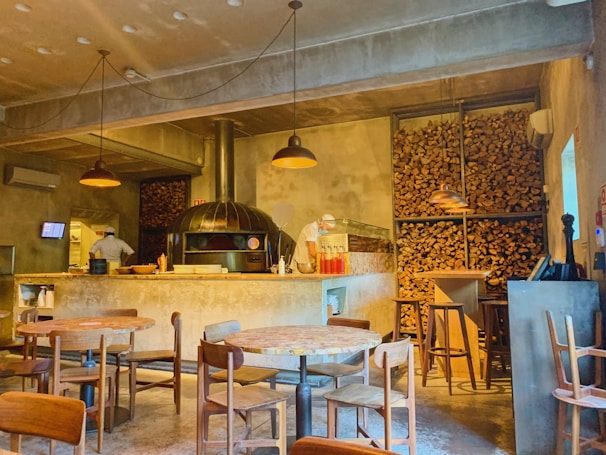 A warmly lit pizzeria with a rustic, industrial design featuring a large wood-fired oven. Stacks of firewood are stored on shelves along the walls. Round wooden tables and chairs are positioned throughout the room. Two chefs are working behind the counter, and there are a few condiments and utensils on top. The concrete floor and ceiling along with pendant lighting add to the cozy atmosphere.