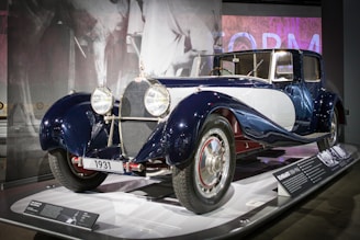 A classic vintage car from 1931 is displayed in a museum exhibit. The car has a sleek and polished exterior with a dark blue and white color scheme. It is equipped with large, circular headlights, chrome accents, and intricate detailing. The background features a monochrome mural of a person and some informational plaques providing details about the car.