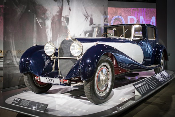 A classic vintage car from 1931 is displayed in a museum exhibit. The car has a sleek and polished exterior with a dark blue and white color scheme. It is equipped with large, circular headlights, chrome accents, and intricate detailing. The background features a monochrome mural of a person and some informational plaques providing details about the car.